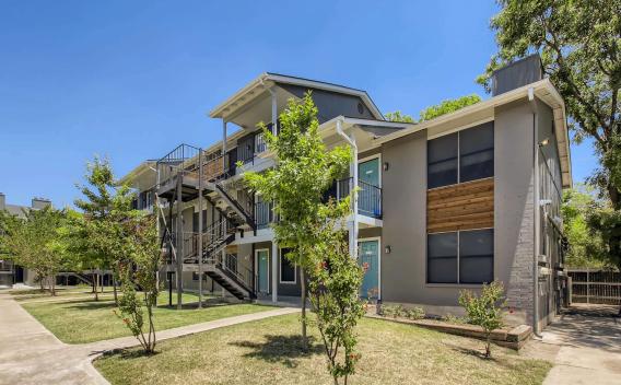 Exterior view of a two-story apartment building with light blue doors and black railings, surrounded by manicured grass and young trees under a clear blue sky.