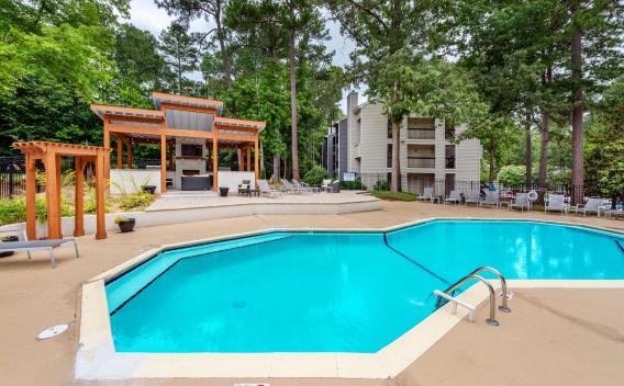 Hexagonal outdoor swimming pool surrounded by lounge chairs, with a modern wooden pergola and grill area set against a lush wooded backdrop and apartment buildings.