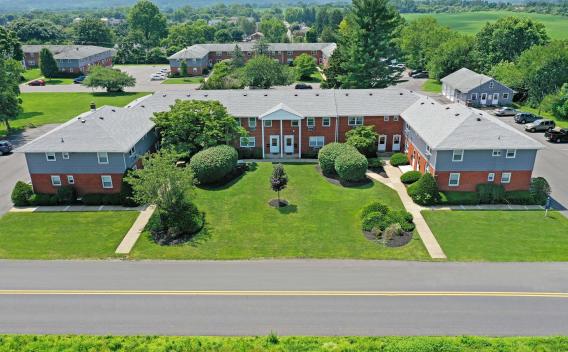 Aerial view of a well-maintained brick apartment building with neatly trimmed landscaping, surrounded by lush greenery and distant rolling hills.