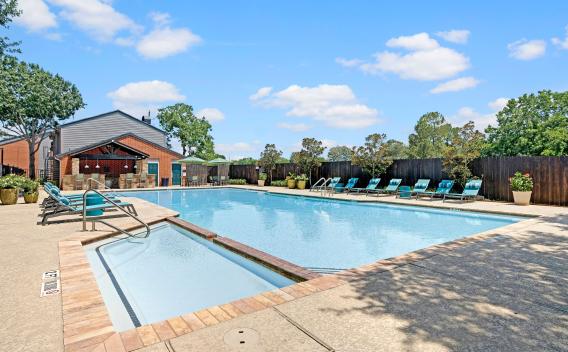 Resort-style swimming pool with poolside lounge chairs, shaded seating area, and a red brick clubhouse under a clear blue sky.