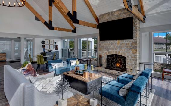 Elegant resident lounge featuring vaulted ceilings with exposed wood beams, a floor-to-ceiling stone fireplace, plush modern seating, and abundant natural light from surrounding windows.