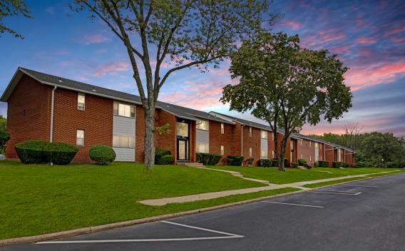The exterior of Laurel Run Village Apartments showcases a well-maintained, two-story brick residential building with manicured green lawns and mature trees. The architecture features a mix of red brick and gray siding, with large windows and dark entrance doors. A spacious parking lot is in the foreground, and a sunset sky in the background adds a warm, inviting atmosphere. ​​