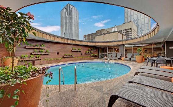 Unique circular rooftop pool surrounded by modern lounge seating and greenery, with views of downtown skyscrapers and urban skyline.