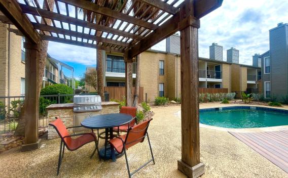 An inviting outdoor amenity space at Oaks of Westchase featuring a shaded pergola with a round dining table and four red chairs. Adjacent to the seating area is a built-in stainless steel grill for outdoor cooking. The area overlooks a sparkling swimming pool, surrounded by lush landscaping and modern apartment buildings with private balconies. The scene is framed by tall palm trees, creating a relaxed and resort-like atmosphere.