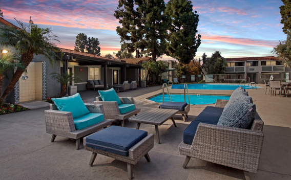 A resort-style swimming pool area at Olive Ridge Apartments captured during twilight. The space features woven wicker lounge chairs with plush blue and turquoise cushions, a communal seating area with ottomans, and a shimmering pool surrounded by lush landscaping. The evening sky is painted in shades of pink and blue, creating a serene atmosphere.