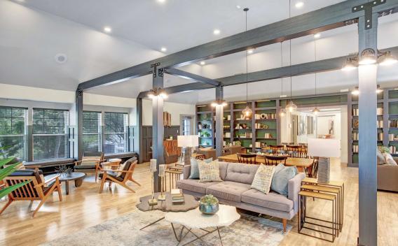 Spacious resident clubhouse with high ceilings, exposed architectural beams, and a warm, inviting design. The room features multiple lounge areas with modern furnishings, a library wall with built-in bookshelves, large windows with natural light, and contemporary pendant lighting. Ideal for social gatherings or relaxing in style.