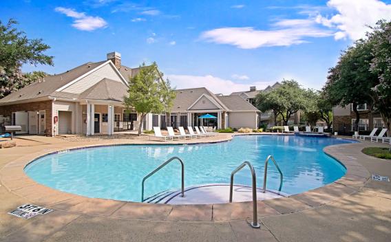 Resort-style swimming pool surrounded by lounge chairs and trees, with a view of the clubhouse and residential buildings in the background under a clear blue sky