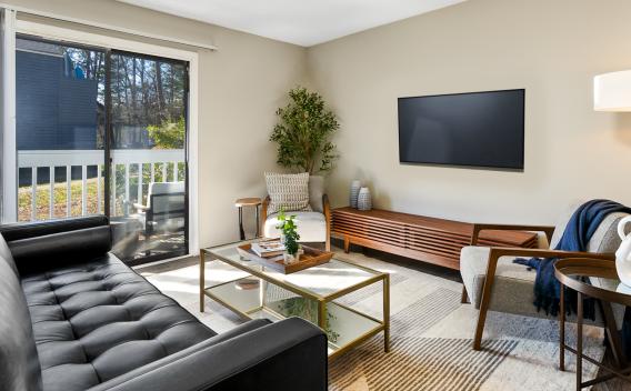 Modern living room at The Forest Apartments featuring a black leather sofa, a glass coffee table with a gold frame, and a cozy armchair with a throw blanket. A large sliding glass door leads to a private balcony with outdoor seating, allowing natural light to fill the space. The room is decorated with neutral tones, a sleek media console, and stylish accents, creating a warm and inviting atmosphere.