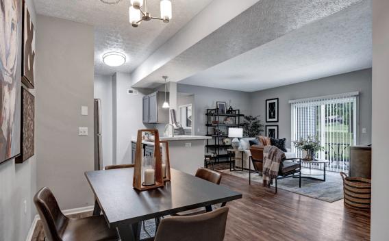 A modern open-concept living space at Trotters Landing featuring a dining area, kitchen, and living room. The dining area has a dark wood table with four leather chairs and decorative candle lanterns. The kitchen includes gray cabinetry, stainless steel appliances, and a white island with pendant lighting. The living room is furnished with a cozy sofa, accent chairs, and a stylish bookshelf. Large sliding glass doors lead to a balcony with a green lawn view, allowing natural light to fill the space. ​​