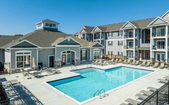 Bright and spacious outdoor pool area with lounge chairs and patio tables, set against the backdrop of the clubhouse and apartment buildings.