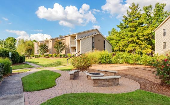 A beautifully landscaped outdoor space at Nova Chapel Hill featuring a circular fire pit with built-in stone seating, surrounded by lush greenery and manicured bushes. A winding pathway leads to the beige-toned apartment buildings with private balconies, nestled among mature trees. The bright blue sky with fluffy white clouds enhances the inviting and peaceful atmosphere of the community.
