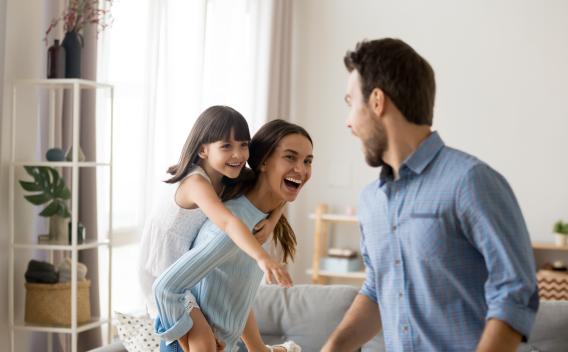 two parents and a girl child in a living room with the mother giving the child a piggyback ride