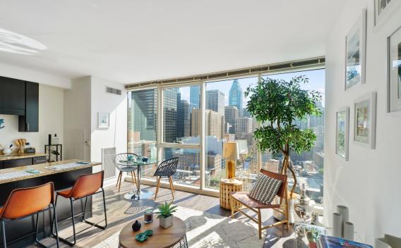 A modern, sunlit apartment living area with floor-to-ceiling windows offering a panoramic view of a city skyline with tall buildings. The room features a cozy seating arrangement including a wooden chair with a patterned cushion, a small round coffee table with decorative items, and a large potted tree near the window. There is a dining area with two leather chairs at a counter and a small round table with two chairs by the window. The space is decorated with framed artwork on the wall, a neutral rug, and c