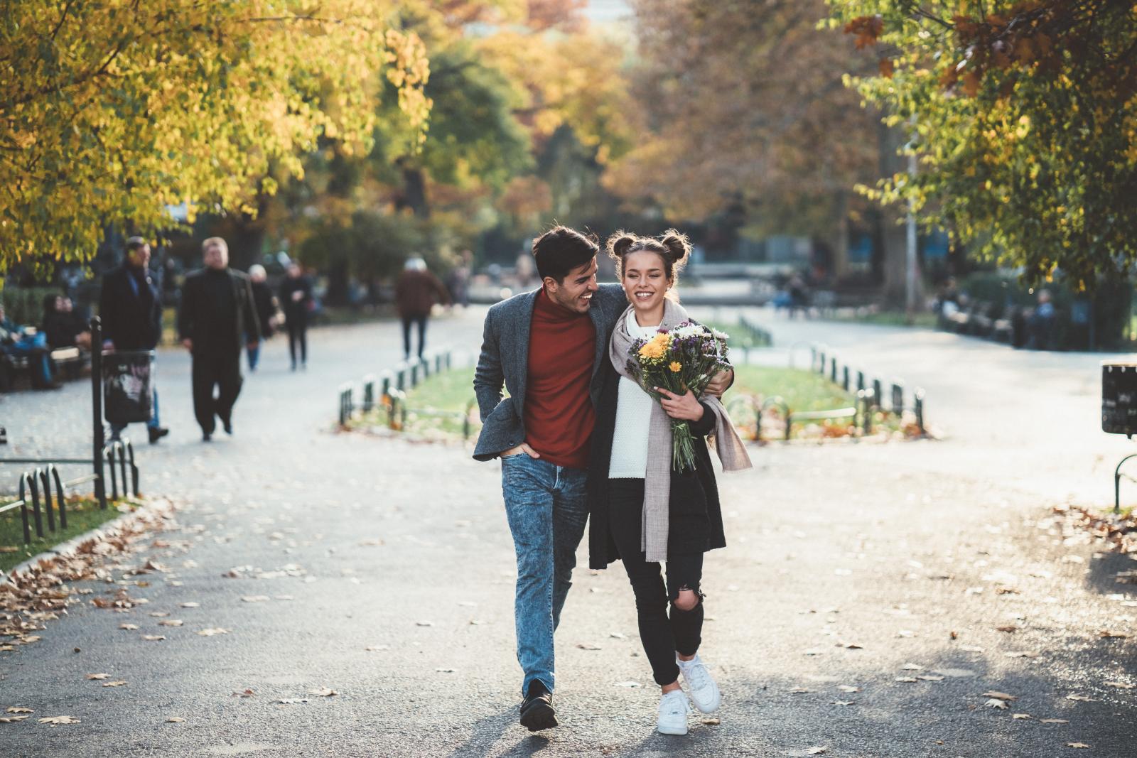 A couple walking arm-in-arm through a park in autumn, with one holding a bouquet of flowers.