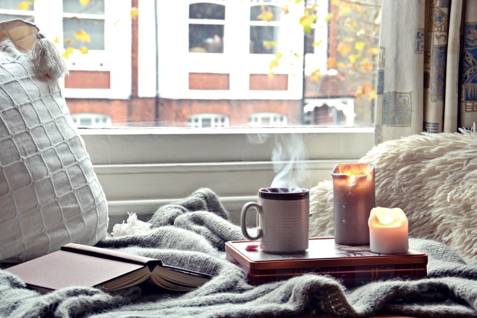 A cozy scene of a hot mug, some candles, and a book by a window overlooking a street.