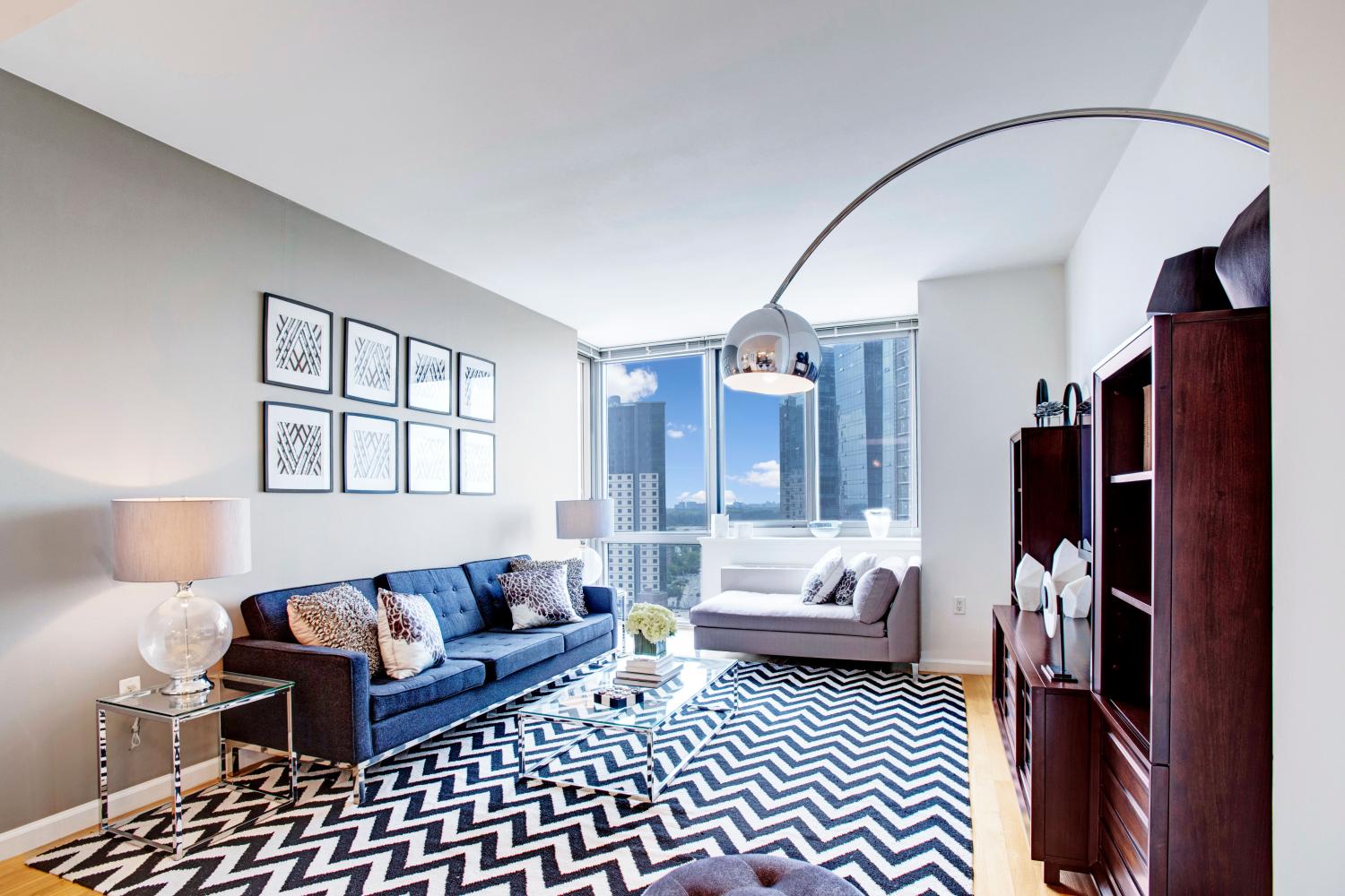 A modern living room with large windows offering city views, stylish furniture, and a bold zigzag patterned rug, creating a vibrant and cozy ambiance.
