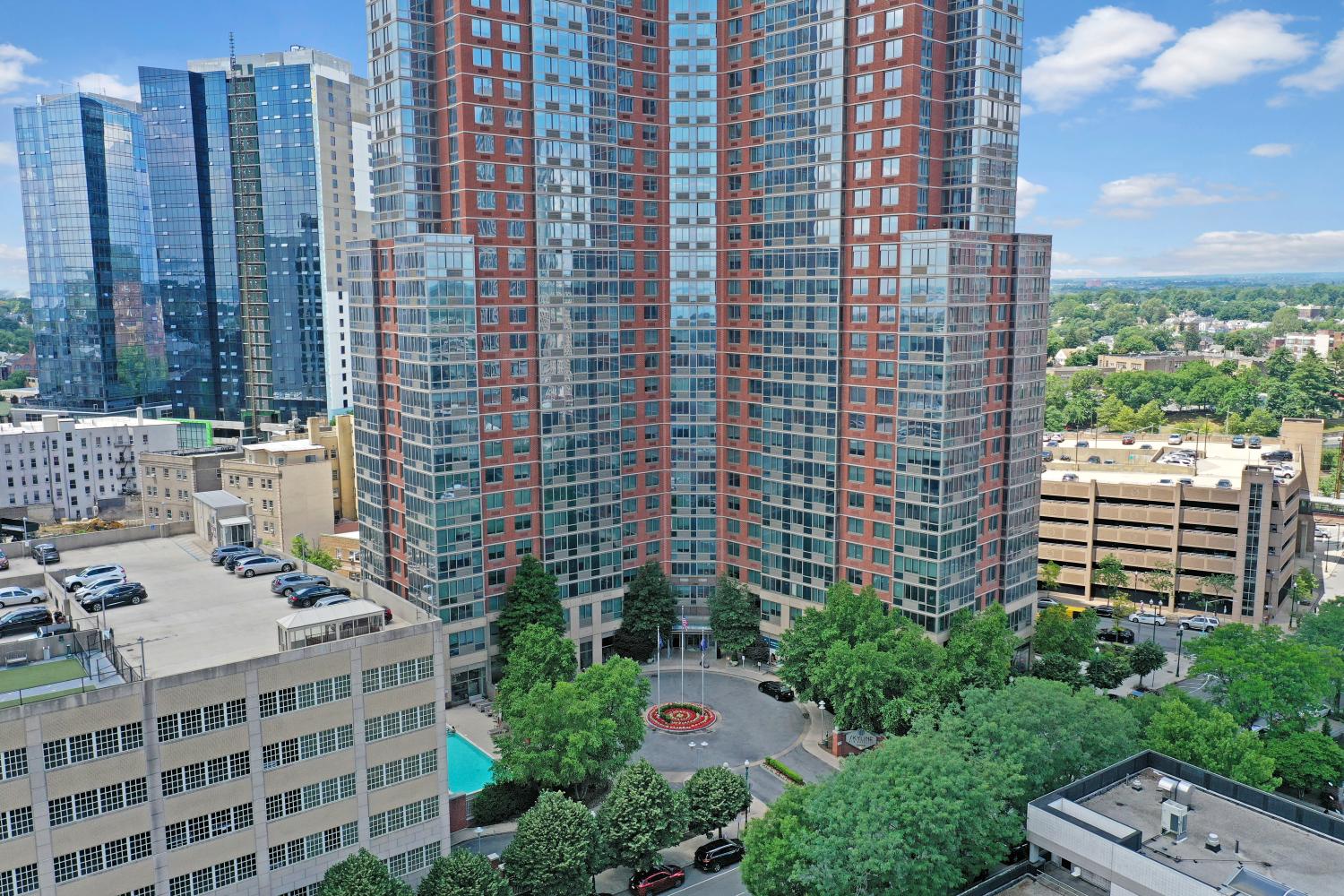 An aerial view showcasing the towering red-brick and glass Skyline New Rochelle apartments surrounded by lush trees and urban scenery.