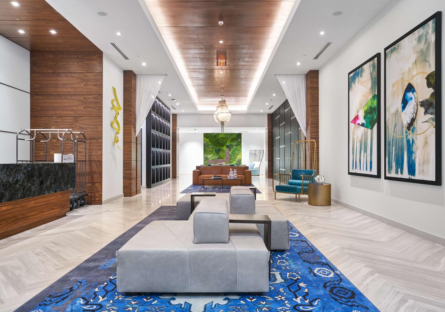Elegant and upscale lobby with a modern design, featuring luxurious lounge seating, bold abstract artwork, and striking architectural elements. The space includes a dramatic wood-paneled ceiling, herringbone marble floors, and a rich blue patterned rug that anchors the room. Tall columns, ambient lighting, and curated decor add to the sophisticated ambiance.