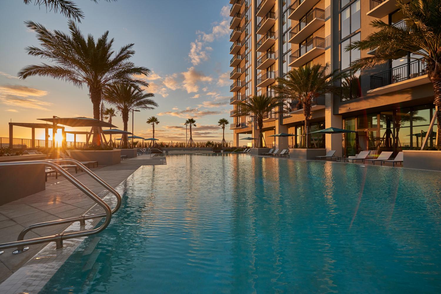 Resort-style pool at Parkline Miami bathed in the warm glow of sunset. Towering palm trees line the tranquil water, while modern loungers and shaded cabanas invite relaxation. The adjacent high-rise reflects the golden hour light, creating a serene and luxurious atmosphere perfect for unwinding at day's end.
