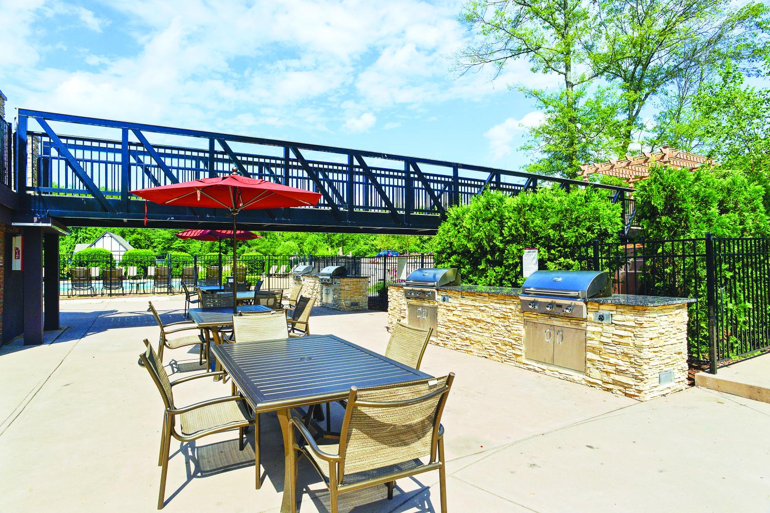 Outdoor grilling and dining area with stainless steel barbecue stations, stone countertops, tables with umbrellas, and a pedestrian bridge overhead, surrounded by greenery.