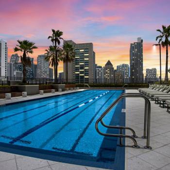 A rooftop lap pool at Parkline Miami Luxury Apartments surrounded by lounge chairs and palm trees, with a breathtaking sunset over the Miami skyline.