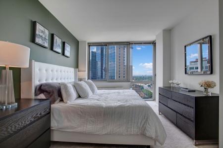 A luxurious bedroom featuring a tufted white headboard, large windows with urban views, and sophisticated decor, providing a tranquil retreat.