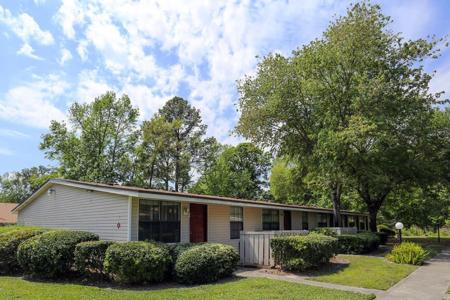 Beige house with a red door and surrounding greenery, set against a backdrop of tall trees and blue sky.