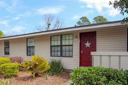 Beige house with a red door adorned with a silver star, surrounded by well-maintained plants and shrubs.