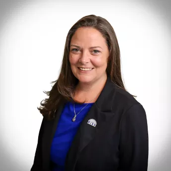 Smiling woman with long brown hair, black blazer, blue top, and silver emblem pin.