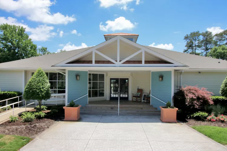 A single-story building with a gabled entrance, blue and white exterior, large windows, and a landscaped walkway leading to double glass doors under a bright blue sky.
