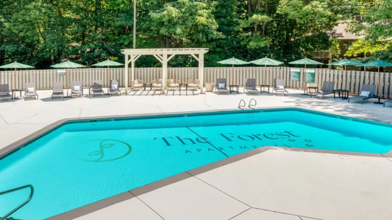  bright outdoor pool area surrounded by lounge chairs, green umbrellas, and a shaded pergola, located at The Forest Apartments, with a backdrop of lush greenery.