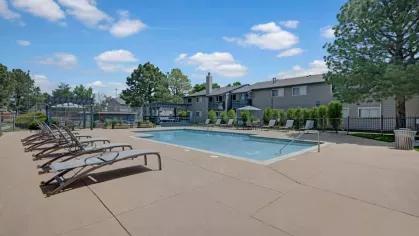  Resort-style swimming pool surrounded by lounge chairs, lush greenery, and modern apartment buildings, offering a relaxing outdoor space under a bright blue sky.