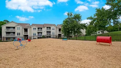 Spacious dog park with agility equipment, open play areas, and a mulch surface, surrounded by modern apartment buildings and lush greenery, providing a fun and safe space for pets.