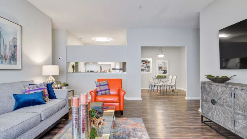 A contemporary apartment interior with an open layout, featuring chic furnishings like a gray sofa, a bold orange armchair, and a glass coffee table, seamlessly connecting to a modern kitchen and dining area.
