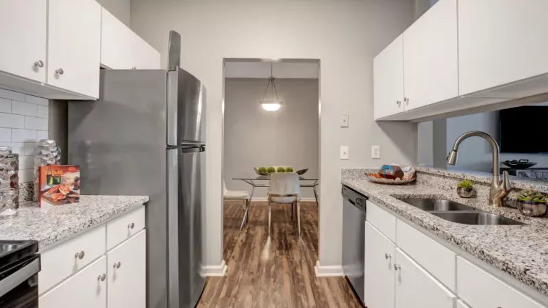 A sleek and modern kitchen featuring granite countertops, stainless steel appliances, and white cabinetry, with a view into a stylish dining area with wood-style flooring and contemporary lighting.