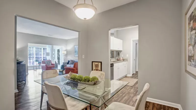 A stylish dining area featuring a glass table with elegant chairs, warm wood-style flooring, and modern lighting, seamlessly connected to both the kitchen and a bright, inviting living space.
