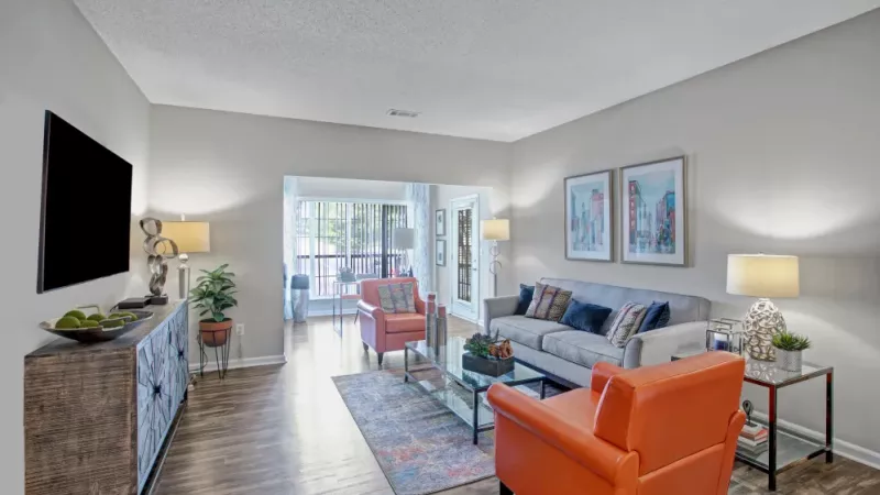 A bright and modern living room featuring a cozy gray sofa, a bold orange armchair, stylish decor, and wood-style flooring, with large windows allowing natural light to fill the space.
