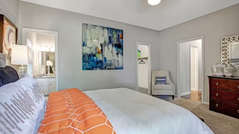 A cozy and modern bedroom featuring a neatly made bed with vibrant orange and white accents, soft carpeting, and stylish decor, including abstract artwork and a decorative mirror, with a walk-in closet and bathroom in view.
