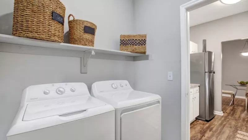 A convenient laundry room featuring a washer and dryer set, a sleek shelf for storage adorned with woven baskets, and a doorway leading to the modern kitchen and dining area.