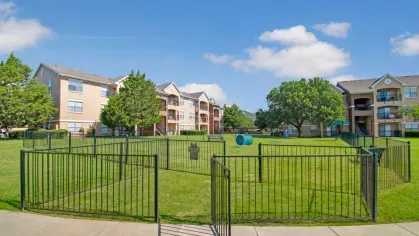 A spacious, fenced dog park with agility equipment surrounded by well-maintained lawns and apartment buildings on a bright, sunny day.