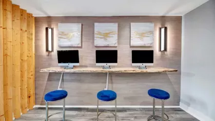 A computer workstation area with three modern monitors on a rustic wooden counter and vibrant blue stools.