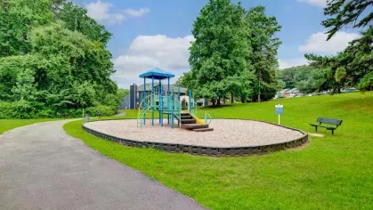 A small playground with a climbing structure and slides set on a circular bed of gravel, surrounded by green lawns and trees.