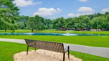 A peaceful bench by a pond with fountains, surrounded by a walking path and lush greenery.