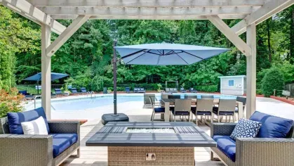 An inviting outdoor seating area with wicker furniture, a fire pit, and a view of the pool, surrounded by lush trees.