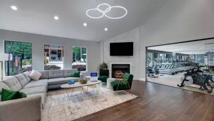 A modern lounge area with a large sectional couch, green armchairs, and a fireplace, adjacent to a gym with glass walls.