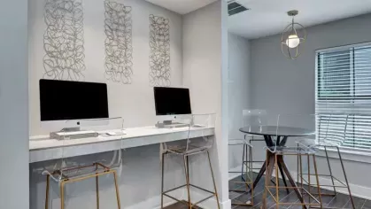 A computer station with two desktop computers and a nearby round table with clear chairs, set in a bright, contemporary space.