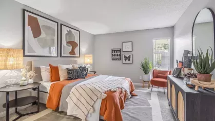 Modern bedroom with earthy tones, featuring a cozy bed with orange accents, abstract wall art, and stylish decor including plants and a full-length mirror.