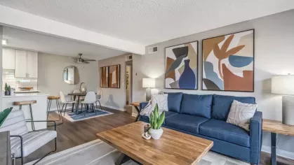 A contemporary living room with a vibrant blue sofa, abstract wall art, wooden furniture, and an open view into the dining and kitchen areas.
