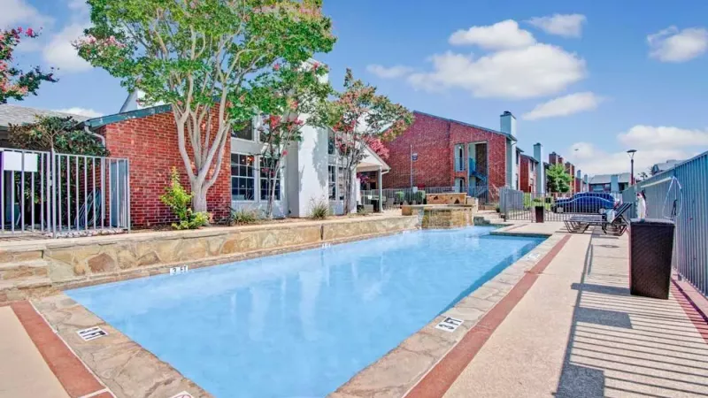 A rectangular swimming pool with a landscaped deck and red brick buildings in the background.