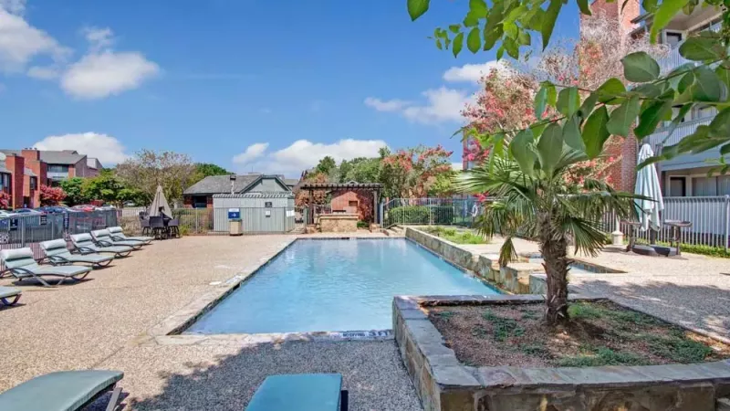 A spacious swimming pool with trees and a small patio area.